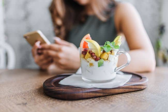 Woman reaching out via phone sitting behind cup of yogurt with fruit